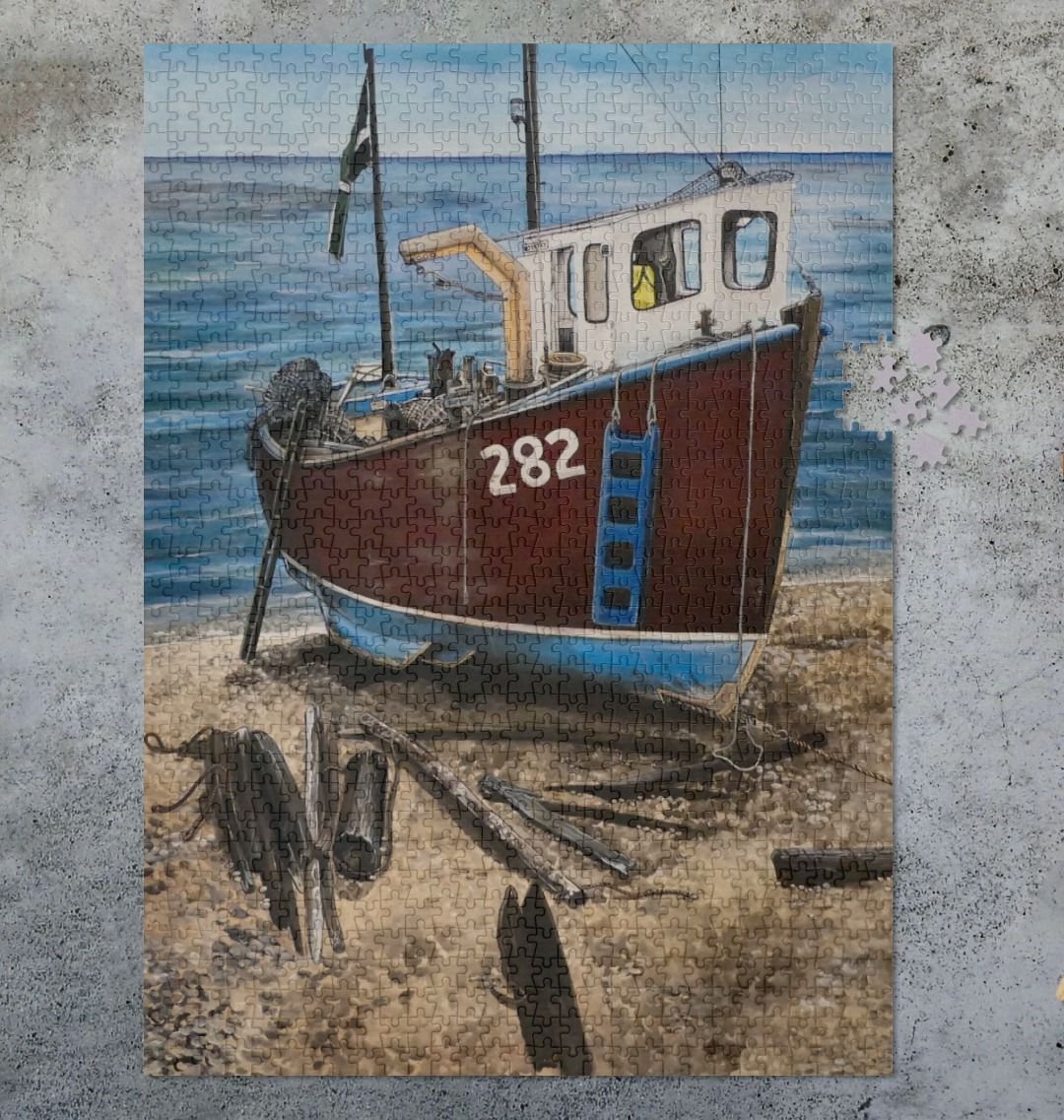 Image of a 1000 Piece Jigsaw Puzzle featuring a copy of the painting 'Relaxing Beer' by Nick Dale. As displayed partially completed on a grey surface. The jigsaw depicts a fishing boat beached at Beer, Devon. The wood is laid under the boats to aid in their winching up the shingle beach.