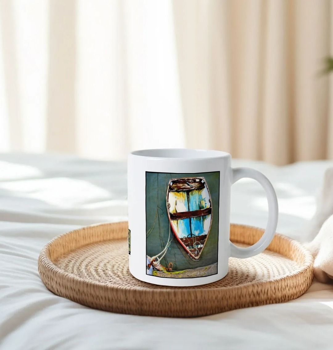 Image of a White Mug on a tray on a bed featuring a copy of the painting 'Good Intentions at Lyme Regis' by Nick Dale. The handle is orientated to the right. The mug depicts a small wooden rowing boat tied up in Lyme Regis harbour, Dorset.