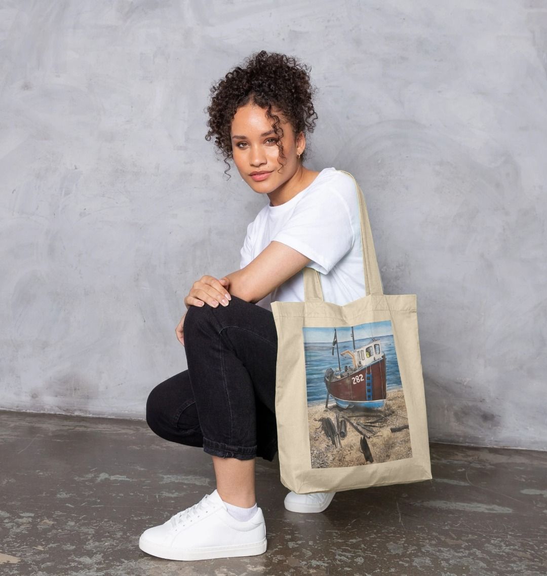 Image of a female model carrying a Natural Tote Bag featuring a copy of the painting 'Relaxing Beer' by Nick Dale. The bag depicts a fishing boat beached at Beer, Devon. The wood is laid under the boats to aid in their winching up the shingle beach.