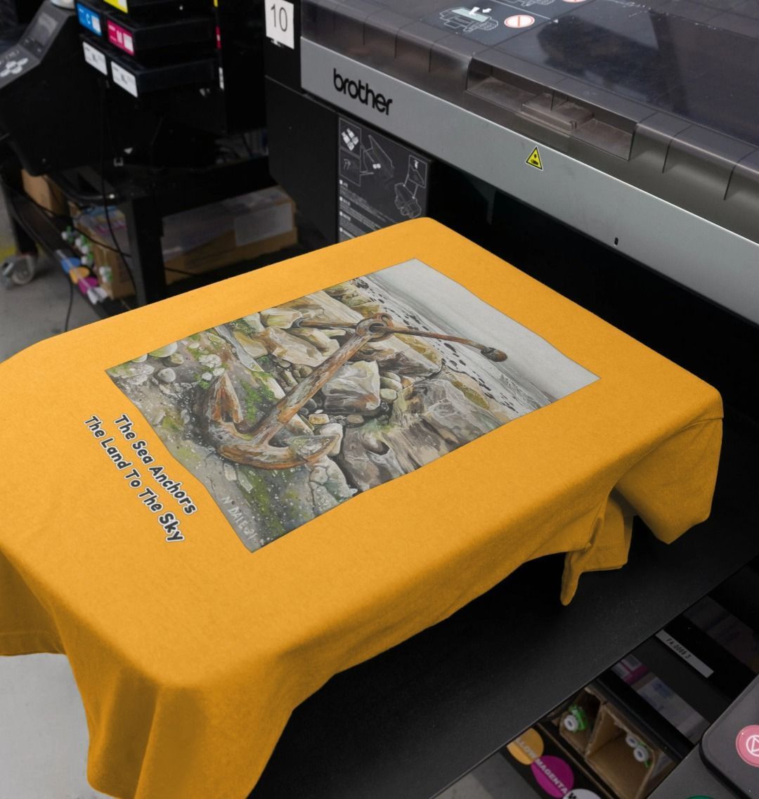 Image of a Mustard Women's Scoop Neck T-shirt featuring a copy of the painting 'The Sea Anchors The Land To The Sky' by Nick Dale, with the words 'The Sea Anchors The Land To The Sky' written beneath it. As shown on a printing machine. The T-shirt depicts an old anchor lying at rest under a leaden sky upon the foreshore at Chapman's Pool, near Worth Matravers, Dorset.