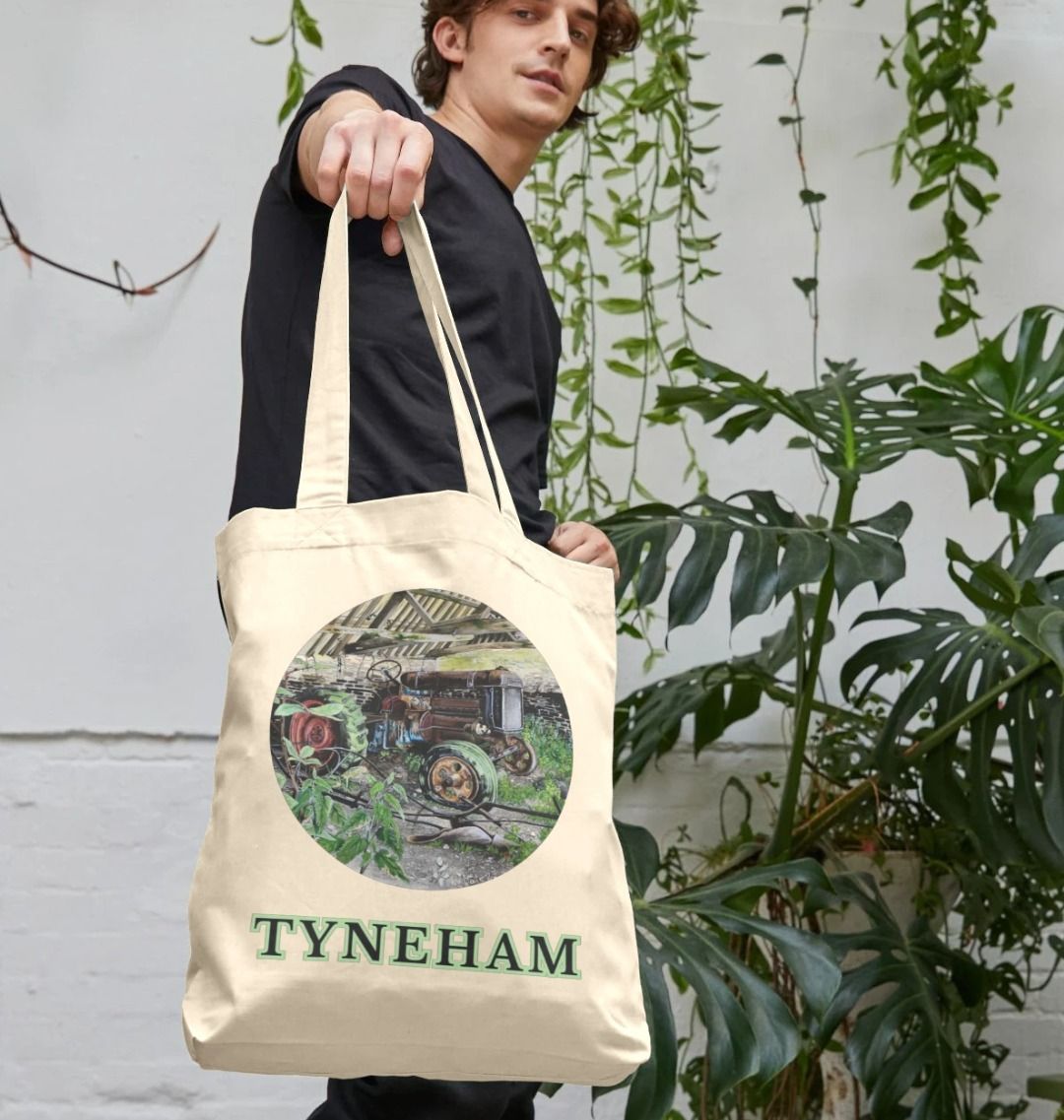 Image of a male model holding a Natural Tote Bag featuring a copy of the painting 'A Well Deserved Rest At Tyneham' by Nick Dale. The bag depicts a Fordson Major tractor laid up in a farm building at the abandoned village of Tyneham in Dorset.