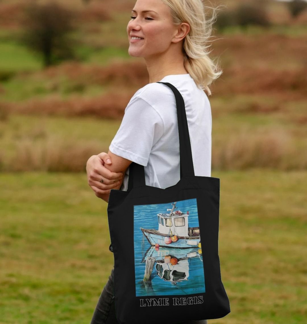 Image of a female model carrying a Black Tote Bag featuring a copy of the painting 'God's Own 99 Flake Pierces The Veil' by Nick Dale, with the words 'Lyme Regis' written beneath it. The bag depicts a fishing boat at rest within the harbour of Lyme Regis, Dorset. The title is based on the similarity of the wooden pile to a chocolate flake as inserted into an ice cream cone. This seemed appropriate given the draw of the town to tourists.