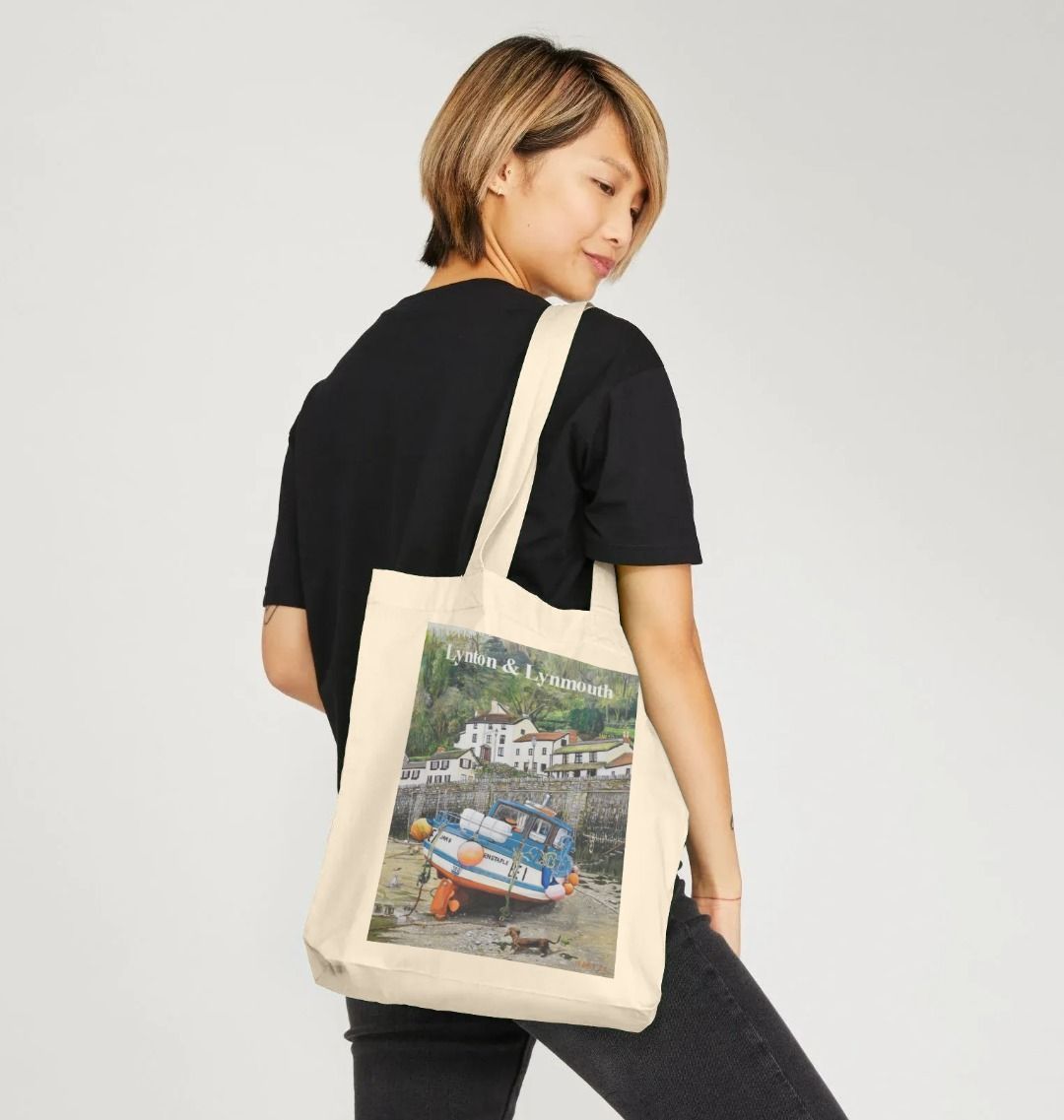 Image of a female model carrying a Natural Tote Bag featuring a copy of the painting 'Lynmouth At Lowest Tide' by Nick Dale, with the words 'Lynton & Lynmouth' written across the top. The bag depicts an unusual perspective on the picturesque harbour of Lynmouth (Devon), as seen from the floor of the harbour at low tide. Behind the fishing boat can be seen the characterful harbourside buildings.