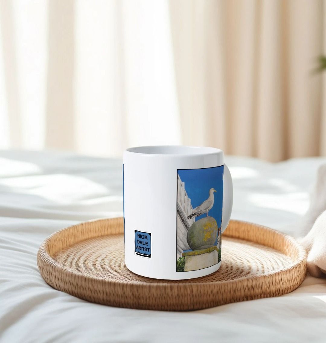 Image of a White Mug on a tray on a bed featuring a copy of the painting 'A View Of The Sea At Lyme Regis' by Nick Dale. Image also shows artist's logo on the mug. The mug handle is obscured behind the mug. The mug depicts a gull and buildings along Marine Parade, Lyme Regis.