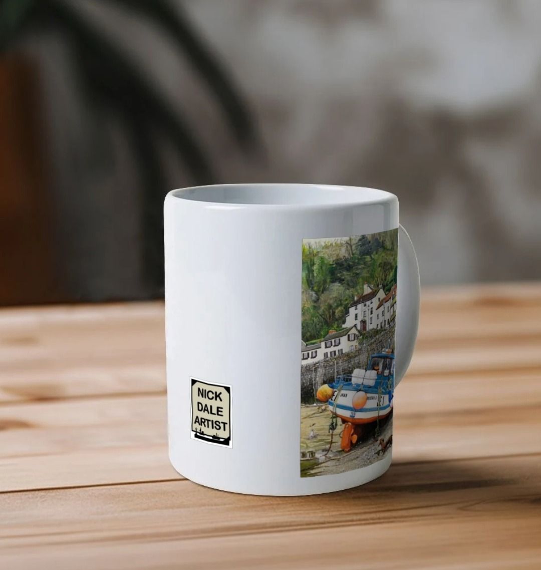 Image of a White Mug on a wooden table featuring a copy of the painting 'Lynmouth At Lowest Tide' by Nick Dale. Image also shows artist's logo on the mug. The mug handle is obscured behind the mug. The mug depicts an unusual perspective on the picturesque harbour of Lynmouth (Devon), as seen from the floor of the harbour at low tide. Behind the fishing boat can be seen the characterful harbourside buildings.