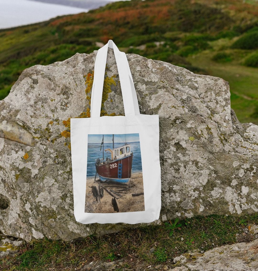 Image of White Tote Bag featuring a copy of the painting 'Relaxing Beer' by Nick Dale. Shown lying on a rock. The bag depicts a fishing boat beached at Beer, Devon. The wood is laid under the boats to aid in their winching up the shingle beach.