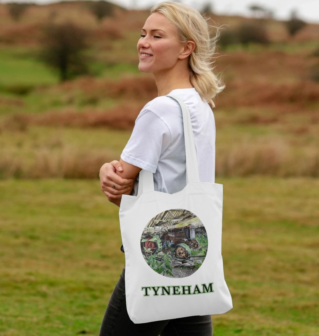 Image of a female model holding a White Tote Bag featuring a copy of the painting 'A Well Deserved Rest At Tyneham' by Nick Dale. The bag depicts a Fordson Major tractor laid up in a farm building at the abandoned village of Tyneham in Dorset.