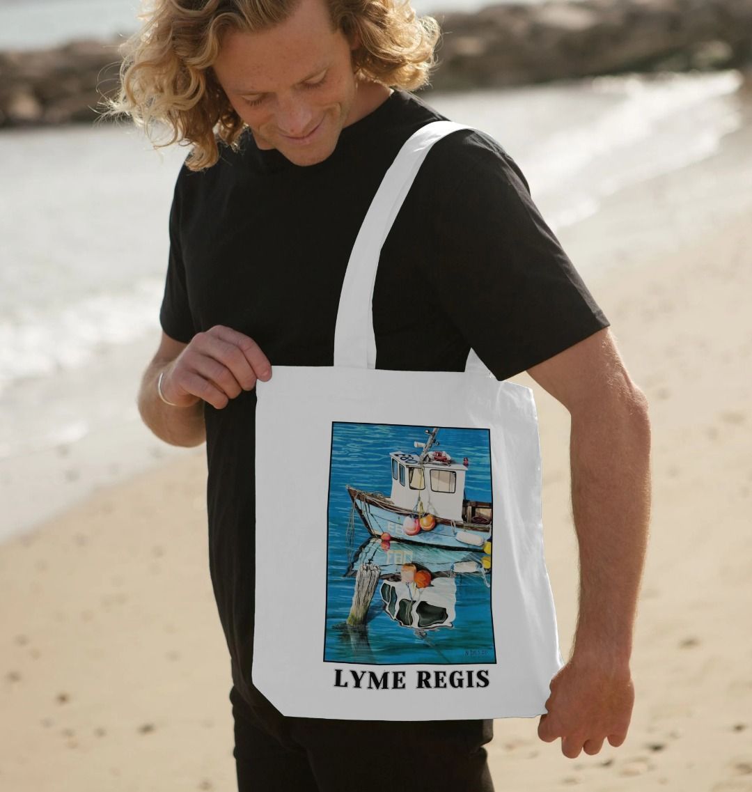 Image of a male model carrying a White Tote Bag featuring a copy of the painting 'God's Own 99 Flake Pierces The Veil' by Nick Dale, with the words 'Lyme Regis' written beneath it. The bag depicts a fishing boat at rest within the harbour of Lyme Regis, Dorset. The title is based on the similarity of the wooden pile to a chocolate flake as inserted into an ice cream cone. This seemed appropriate given the draw of the town to tourists.