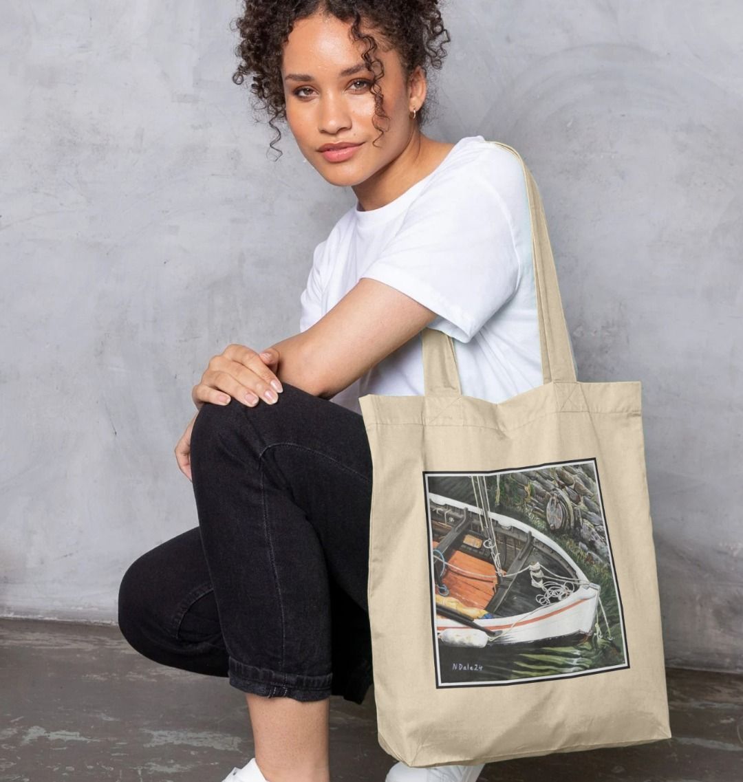 Image of a female model carrying a Natural Tote Bag featuring a copy of the painting 'The Outlet' by Nick Dale. The bag depicts a view down into the harbour at Lynmouth in North Devon showing a small sailing boat at rest against the harbour wall next to a water outfall pipe.