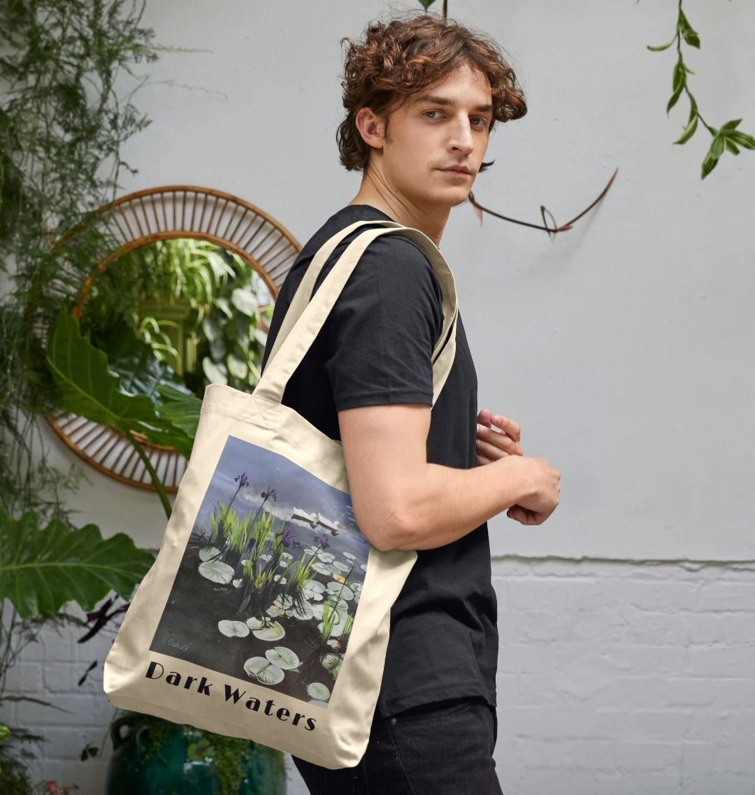 Image of a male model carrying a Natural Tote Bag featuring a copy of the painting 'Dark Waters' by Nick Dale, with the words 'Dark Waters' written beneath it. The bag depicts lily pads, irises and ducks on a pond at Knightshayes Court, Devon.