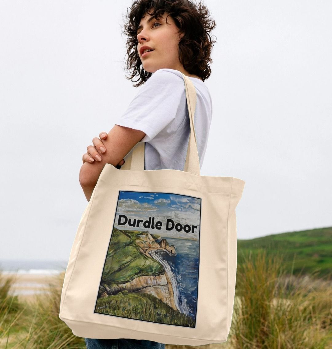 Image of female model carrying a Natural Shopper Tote Bag featuring a copy of the painting 'Durdle Door From The West' by Nick Dale with the words 'Durdle Door' written across it. The bag depicts a view along the Jurassic Coast to the famous Dorset limestone arch landmark, as seen from the coastal path to the west.