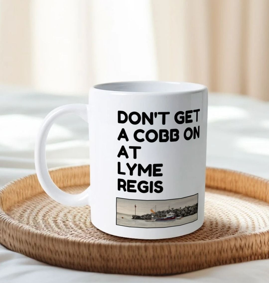 Image of a White Mug on a tray on a bed featuring a copy of the painting 'End Of The Line' by Nick Dale with the slogan 'Don't Get A Cobb On At Lyme Regis' written above it. The handle is orientated to the left. The mug depicts a fisherman preparing his catch for unloading at Lyme Regis as the birds line up on the breakwater and patiently await their turn to feed.