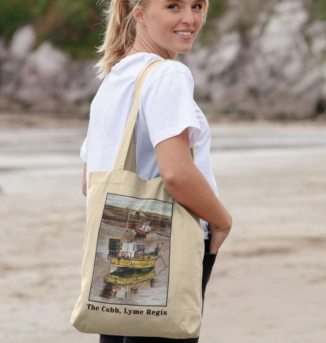 Image of a female model carrying a Natural Tote Bag featuring a copy of the painting 'Lyme Regis at Low Tide' by Nick Dale, with the words 'The Cobb, Lyme Regis' written beneath it. The bag depicts a sunset where the fishing vessels rest on the floor of Lyme Regis harbour whilst the tourists enjoy the view from the Cobb.