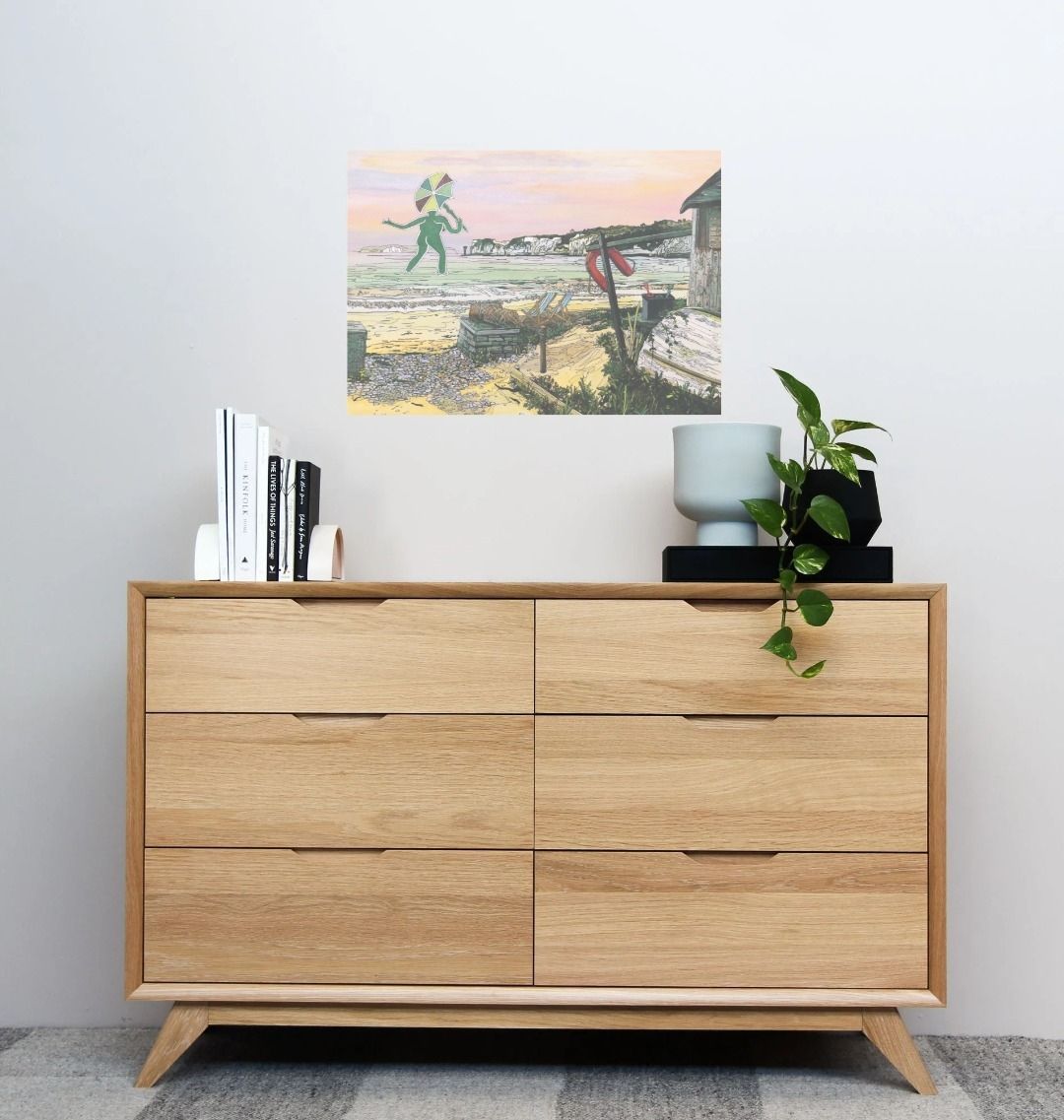 Image of a Large Art Print featuring a copy of the painting 'Mrs Cerne Abbas Giant Visits Studland' by Nick Dale; as displayed on a wall above a chest of drawers. The print imagines the Cerne Abbas Giant's wife visiting the seaside at Studland Beach; with Old Harry Rocks and the Isle of Wight in the distance.