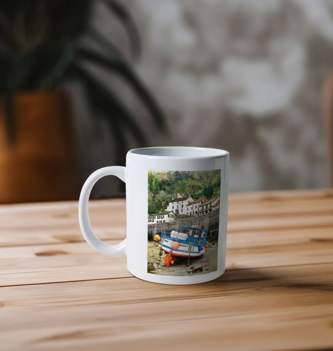 Image of a White Mug on a wooden table featuring a copy of the painting 'Lynmouth At Lowest Tide' by Nick Dale. The handle is orientated to the left. The mug depicts an unusual perspective on the picturesque harbour of Lynmouth (Devon), as seen from the floor of the harbour at low tide. Behind the fishing boat can be seen the characterful harbourside buildings.