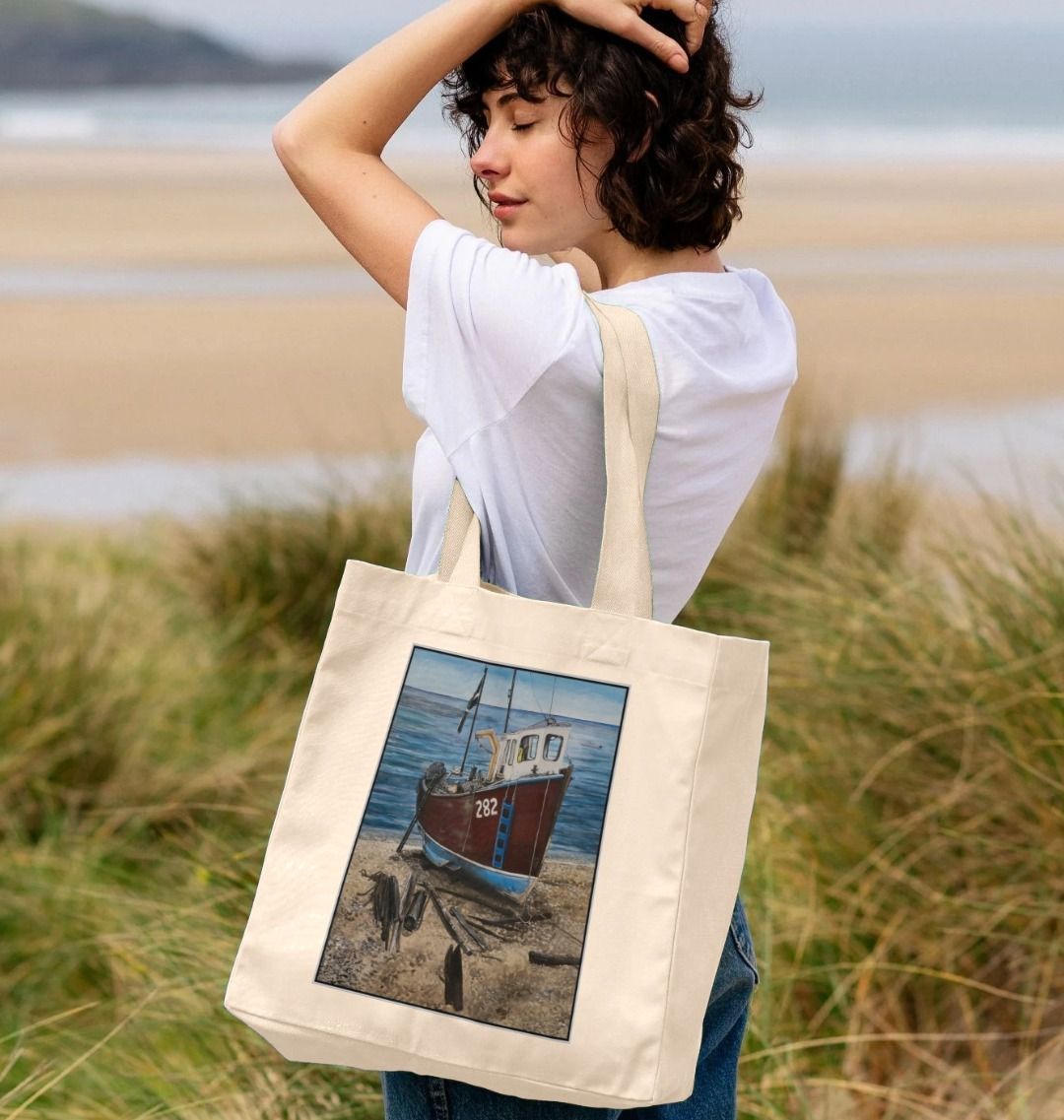 Image of female model carrying a Natural Shopper Tote Bag featuring a copy of the painting 'Relaxing Beer' by Nick Dale. The bag depicts a fishing boat beached at Beer, Devon. The wood is laid under the boats to aid in their winching up the shingle beach.