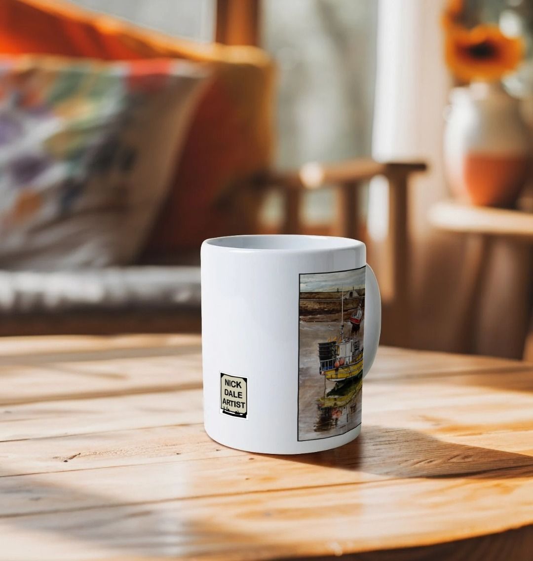 Image of a White Mug on a wooden table featuring a copy of the painting 'Lyme Regis at Low Tide' by Nick Dale. Image also shows artist's logo on the mug. The mug handle is obscured behind the mug. The mug depicts a sunset where the fishing vessels rest on the floor of Lyme Regis harbour whilst the tourists enjoy the view from the Cobb.
