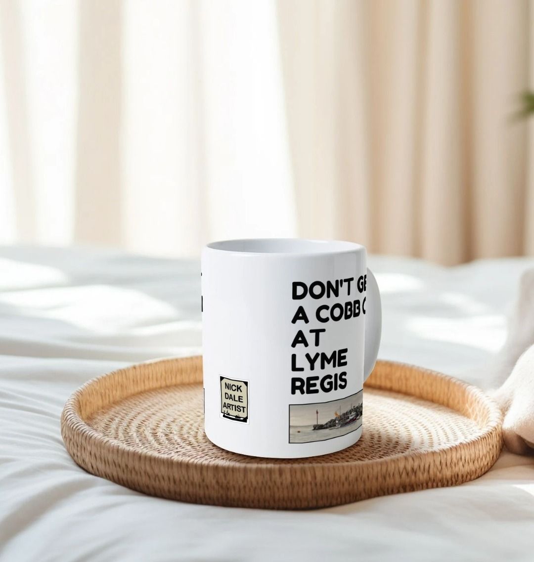 Image of a White Mug on a tray on a bed featuring a copy of the painting 'End Of The Line' by Nick Dale with the slogan 'Don't Get A Cobb On At Lyme Regis' written above it. Image also shows the artist's logo on the mug. The mug handle is obscured behind the mug. The mug depicts a fisherman preparing his catch for unloading at Lyme Regis as the birds line up on the breakwater and patiently await their turn to feed.